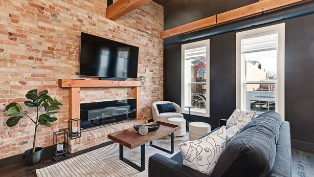 Una sala de estar con pared de ladrillo, un televisor de pantalla plana montado sobre una chimenea, un sofá, una mesa de centro y un ventanal con vistas a un edificio.