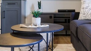 A modern living room with a grey sofa, a round coffee table with a plant, a flat-screen TV mounted on the wall, and a small cabinet.