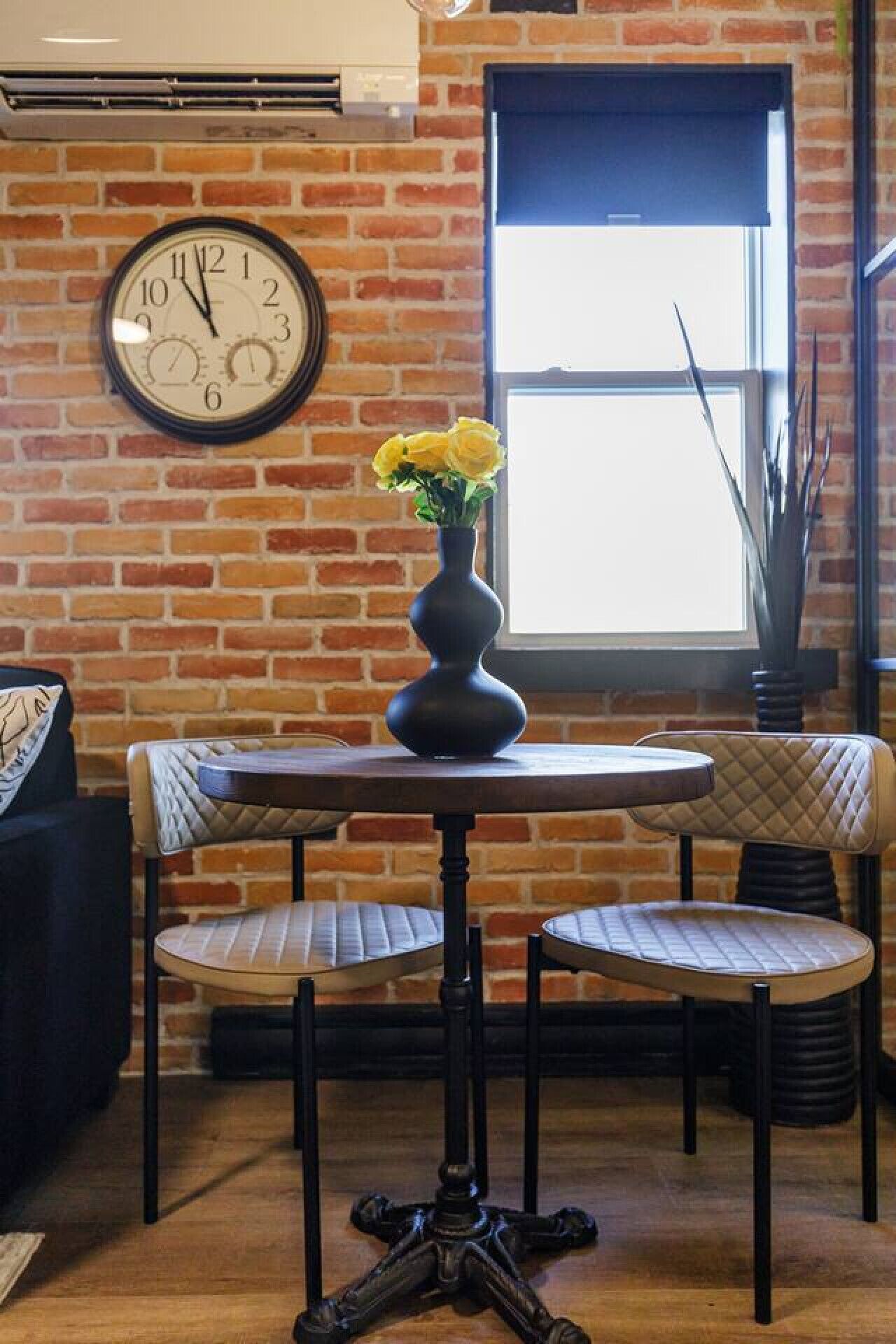 A brick wall with a large analog clock, a round table with two chairs, and a vase with yellow flowers.