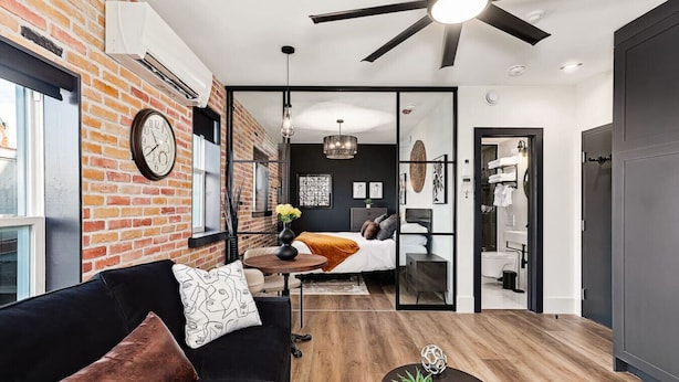 A modern apartment with a brick wall, a black leather sofa, a wooden floor, and a ceiling fan.