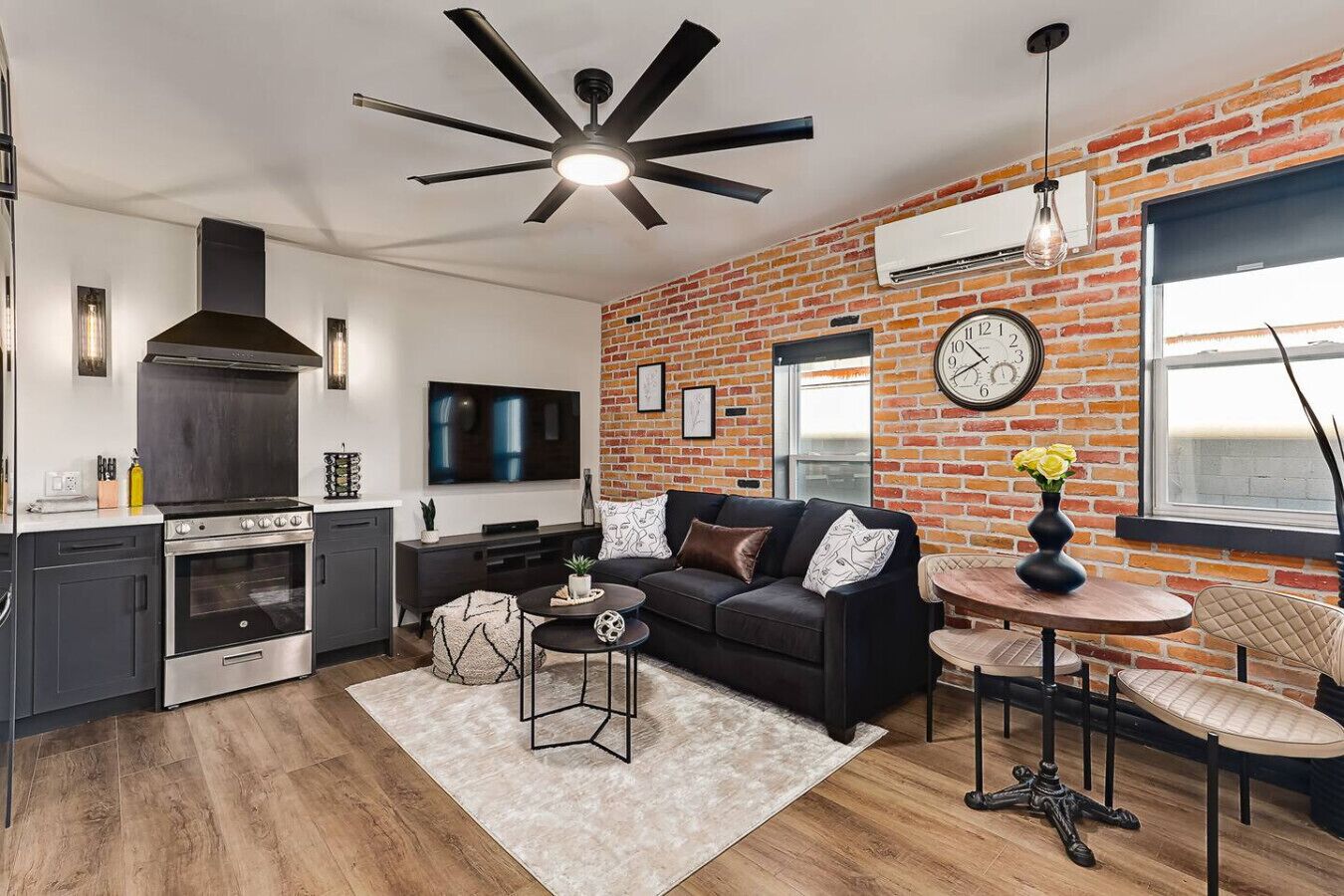 A modern kitchen with a built-in oven and a large TV. There is a black sofa with a coffee table and a small side table with a vase of flowers.