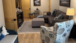 Una sala de estar con sofá, reposapiés y sillón, piso de madera y dos cuadros en las paredes.