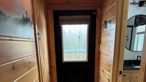 Interior de una cabaña de madera con una puerta que da a un balcón, una alfombra de bienvenida y vistas a árboles por la ventana.