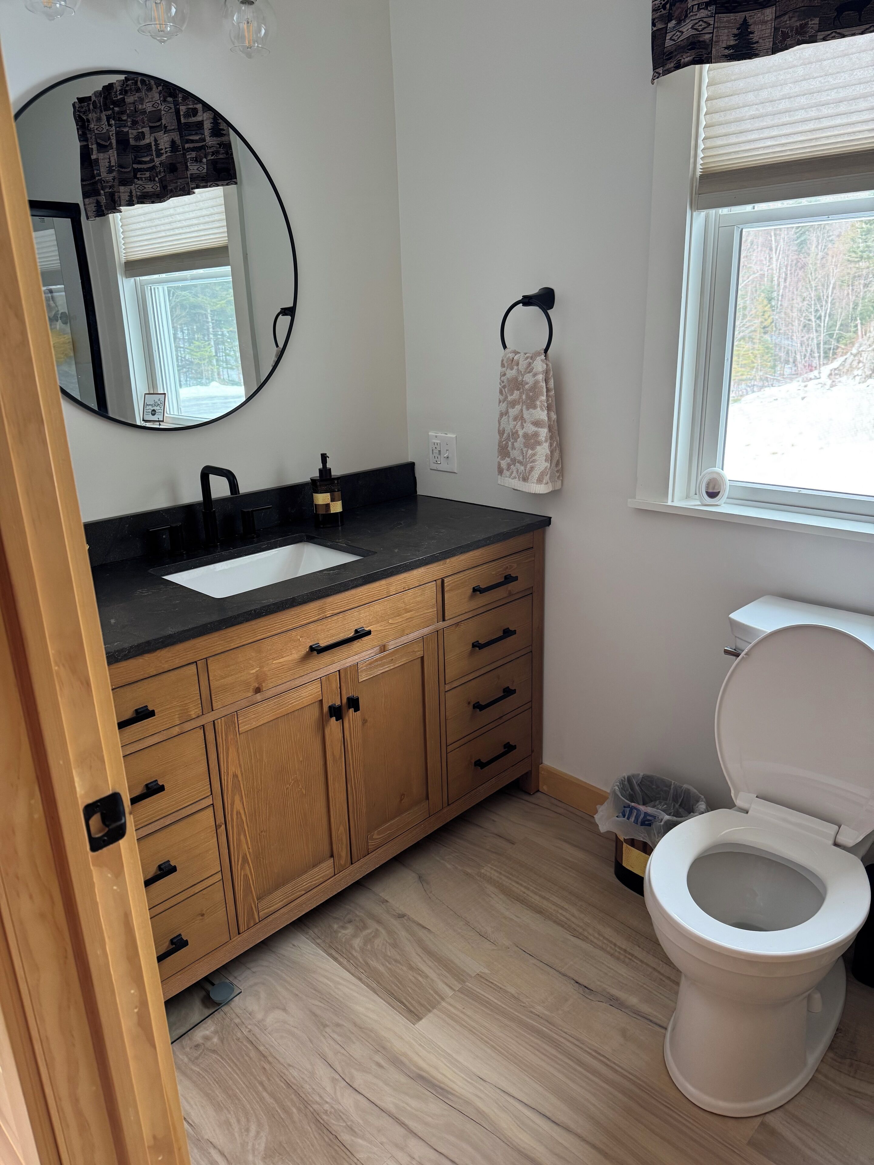 A bathroom with a wooden vanity, a mirror, a toilet, and a window with blinds.