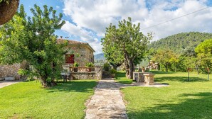 A stone pathway leading to a stone building with a red window, surrounded by lush greenery and a mountainous backdrop. - Can Calistro: Rural Villa with Private Tennis Court (Selva)