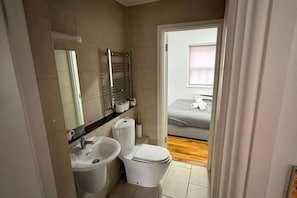 Ein kleines Badezimmer mit Waschbecken, Toilette und Spiegel. Ein Bett ist im angrenzenden Raum zu sehen.