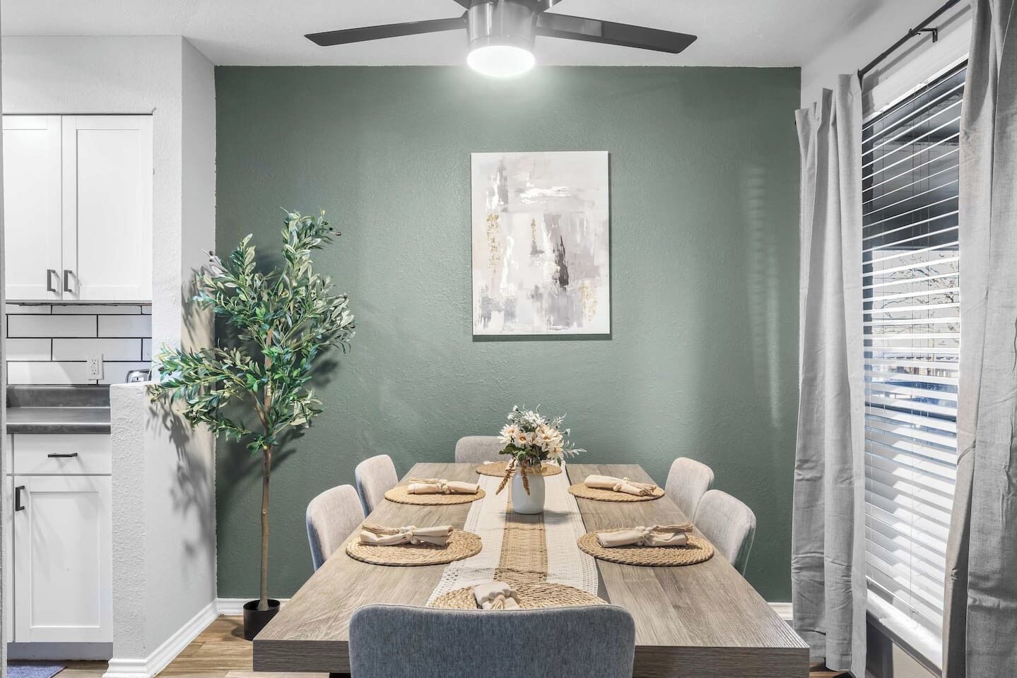 Un comedor con una mesa de madera, sillas, un florero con flores, un cuadro en la pared, un ventilador de techo y una ventana con persianas.