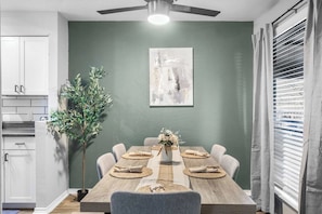 A dining area with a wooden table, chairs, a vase with flowers, a painting on the wall, a ceiling fan, and a window with blinds. - Arlington 3BR | AT&T, Globe Life, 6 Flags, Parking (Arlington)