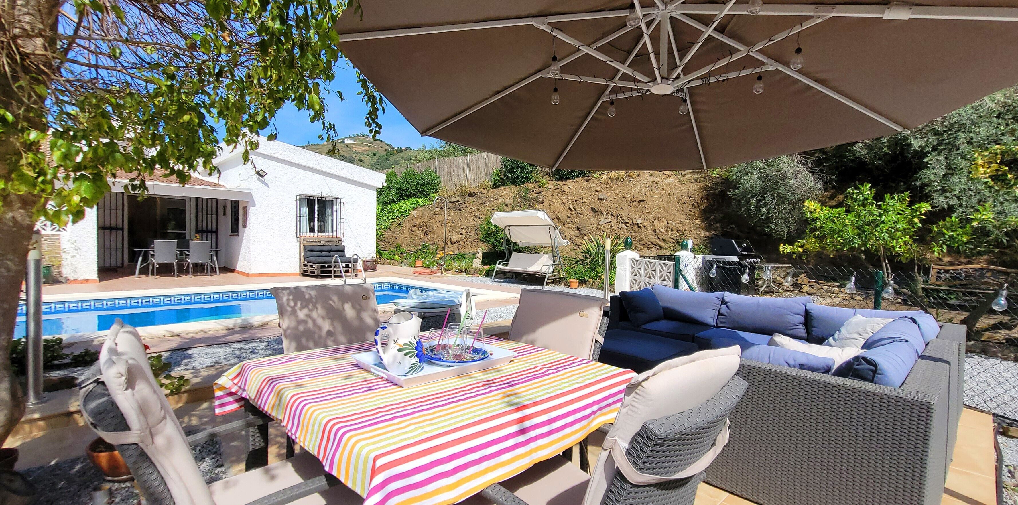 Un patio al aire libre con piscina, una mesa de comedor y un sofá bajo un paraguas grande.