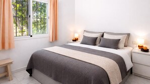 Ein ordentlich bezogenes Bett mit grau-beiger Decke, zwei Nachttischlampen, ein kleiner Holz-Nachttisch und ein Fenster mit Vorhängen.