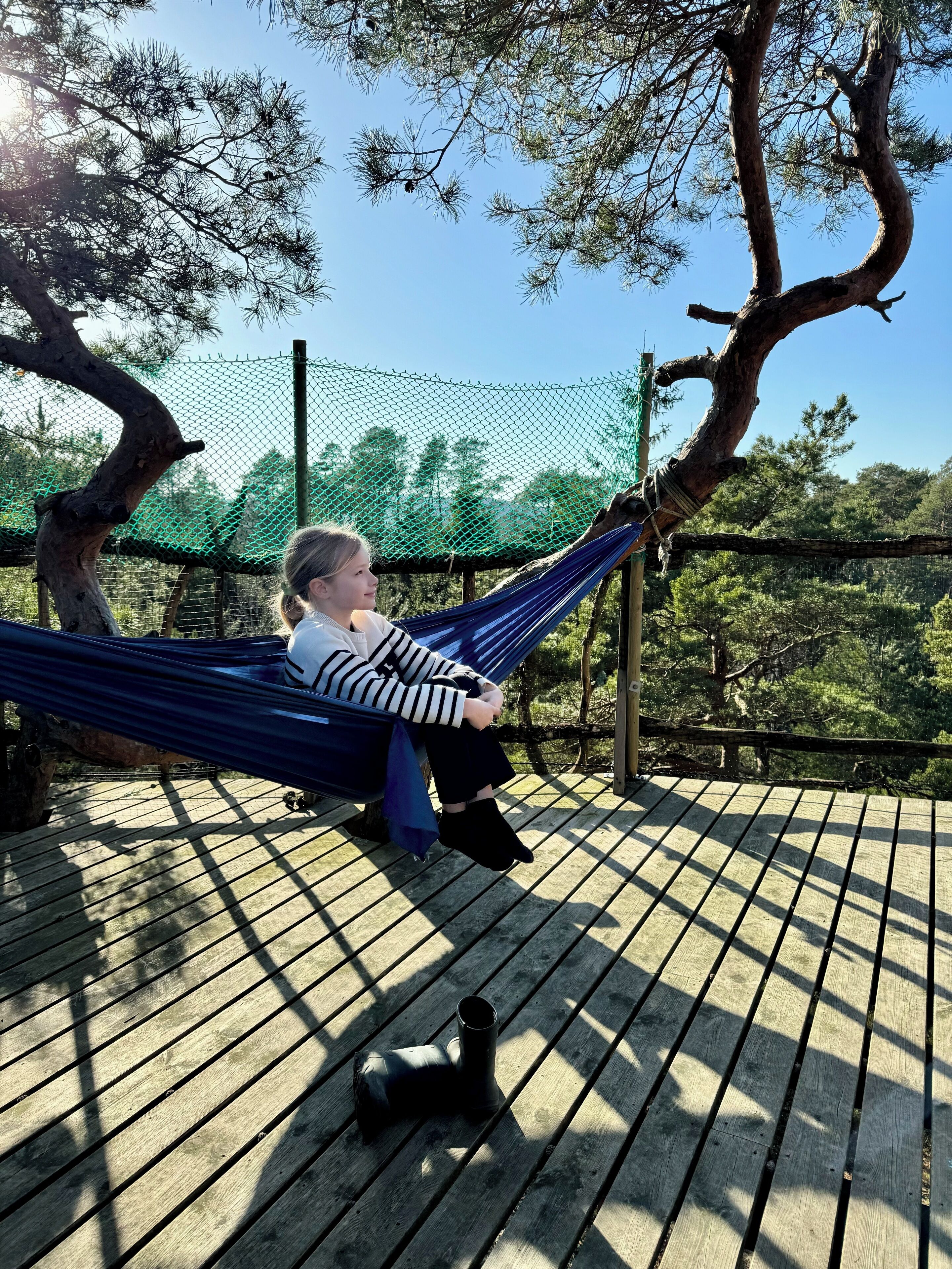 A person relaxing on a hammock on a wooden deck with a view of trees and a fence in the background.