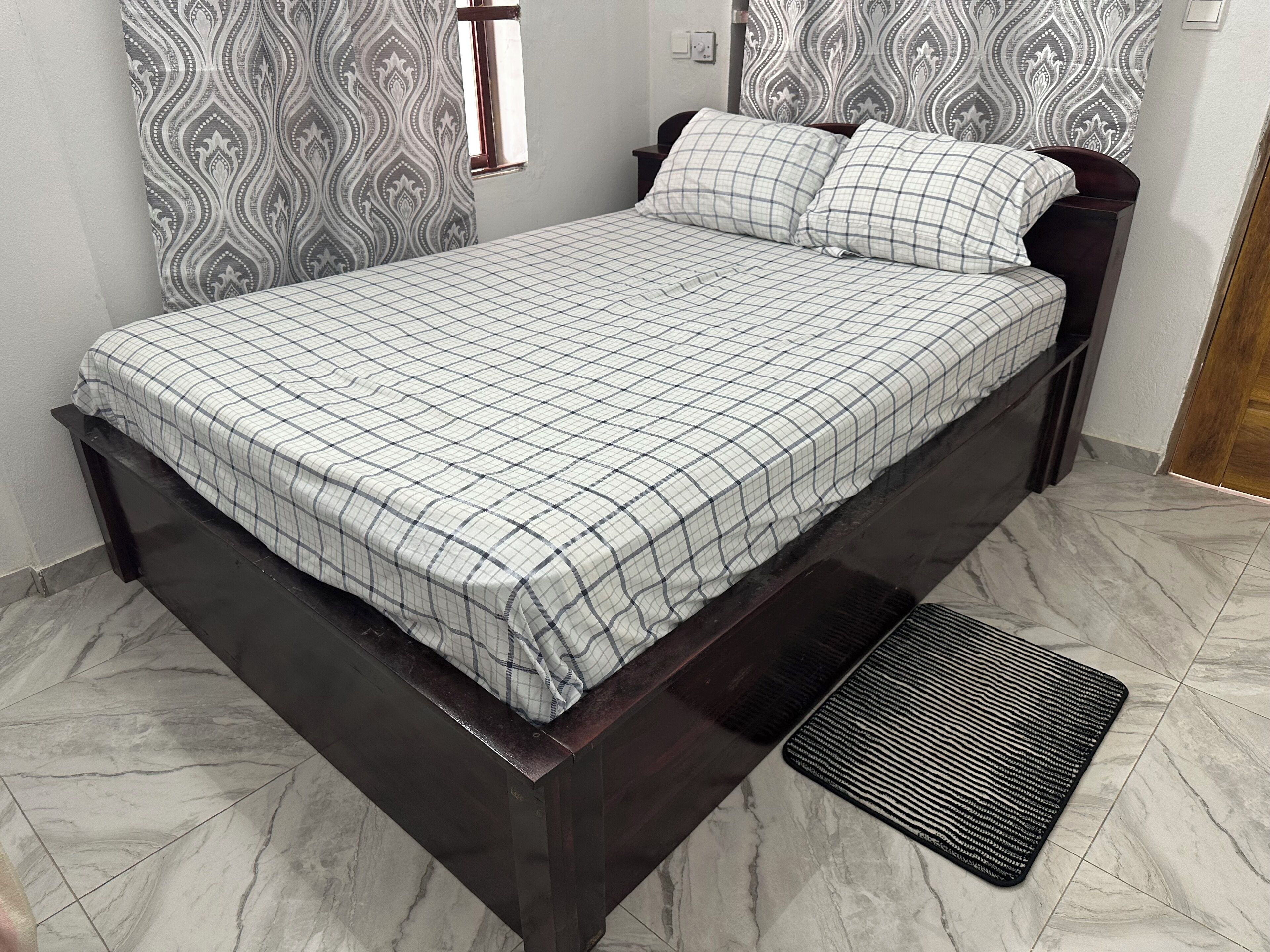 A bed with a checkered bedspread, a dark wooden headboard, and a black textured mat at the foot of the bed.