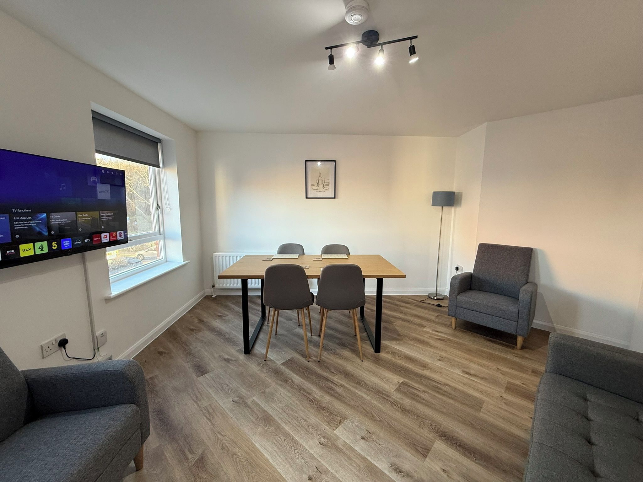 A modern living room with a flat-screen TV, a dining table with four chairs, a grey sofa, and a standing lamp.