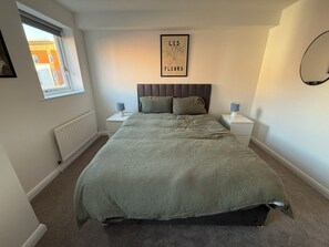 A bedroom with a bed, two bedside tables, a window, a radiator, and a framed picture on the wall. - Modern 2BDR Flat in East London Near Beckton DLR (Greater London)