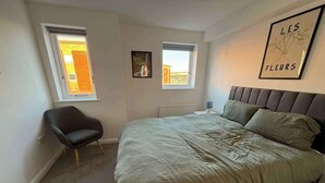 Una camera da letto con un letto, una sedia, due finestre, un termosifone e un quadro appeso al muro.