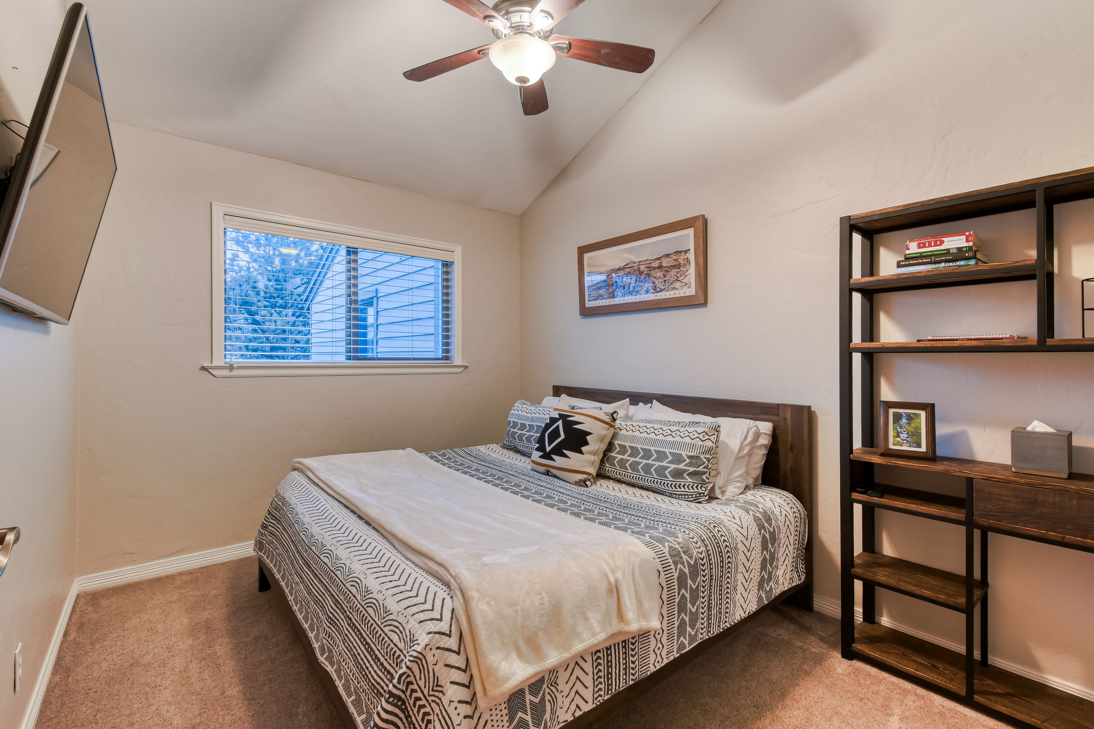 A bedroom with a bed, a ceiling fan, a window, a bookshelf, and a framed picture on the wall.