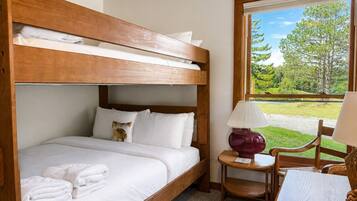 A bunk bed with white bedding, a small table with a lamp, and a view of greenery outside.