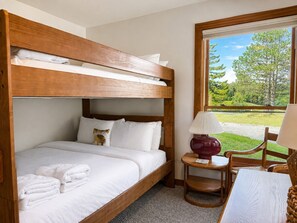 A bunk bed with white bedding, a small table with a lamp, and a view of greenery outside.