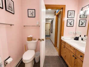 Une salle de bain avec des toilettes, un lavabo et un miroir. Les murs sont ornés de tableaux encadrés et une porte donne accès à une autre pièce.