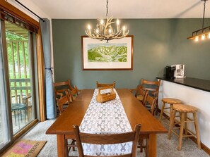 Un coin repas avec une longue table en bois, six chaises, un chemin de table à motifs, un lustre, un tableau représentant un paysage encadré et une machine à café posée sur un comptoir.