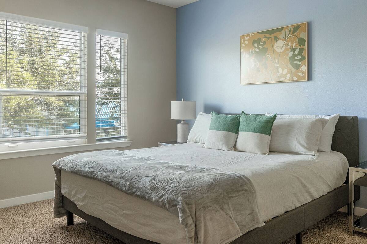 Une chambre à coucher avec un lit, deux tables de chevet avec des lampes, une fenêtre avec des stores et un tableau accroché au mur.