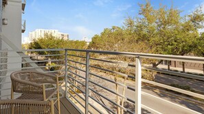 A balcony with wicker chairs, a metal railing, and a view of a street with trees and buildings. - Landing Furnished Apartments - Clearwater (Clearwater)