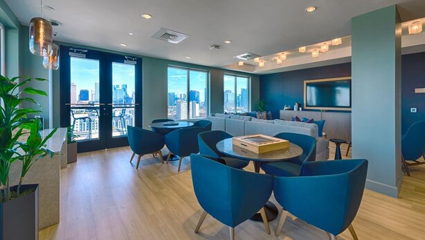 Ein modernes Hotelzimmer mit großen Fenstern, Blick auf die Stadt, einem Essbereich mit blauen Stühlen und einem an der Wand befestigten Flachbildfernseher.