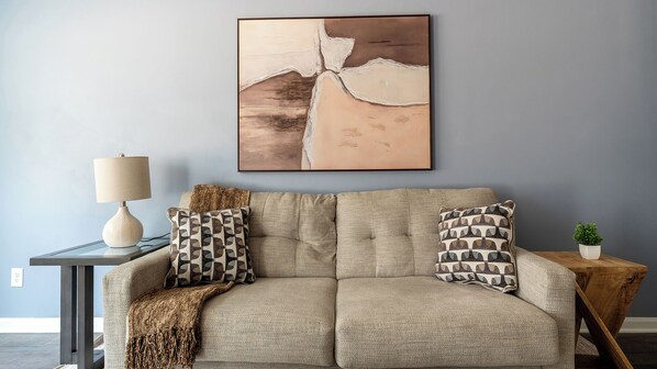 A living room with a sofa, two patterned pillows, a lamp, a framed abstract painting, and a small potted plant. - Landing Furnished Apartments - Bloomingdale (Bloomingdale)