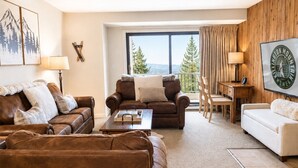 Een woonkamer met een bruine leren bank, een witte bank, een houten salontafel en uitzicht op bomen door een groot raam.