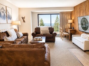A living room with a brown leather sofa, a white sofa, a wooden coffee table, and a view of trees through a large window. - Powder Pad- Pool, Hot tub, Near Bike Park (Killington)