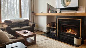 A cozy living room with a fireplace, a sofa, a coffee table, and a television mounted above the fireplace. - Hilltopper Haus- Pool, hot tub, Private Sauna (Killington)