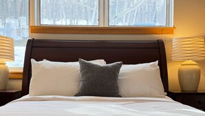 A neatly made bed with white linens and a gray throw blanket, positioned in front of a window with a view of trees. - Hilltopper Haus- Pool, hot tub, Private Sauna (Killington)