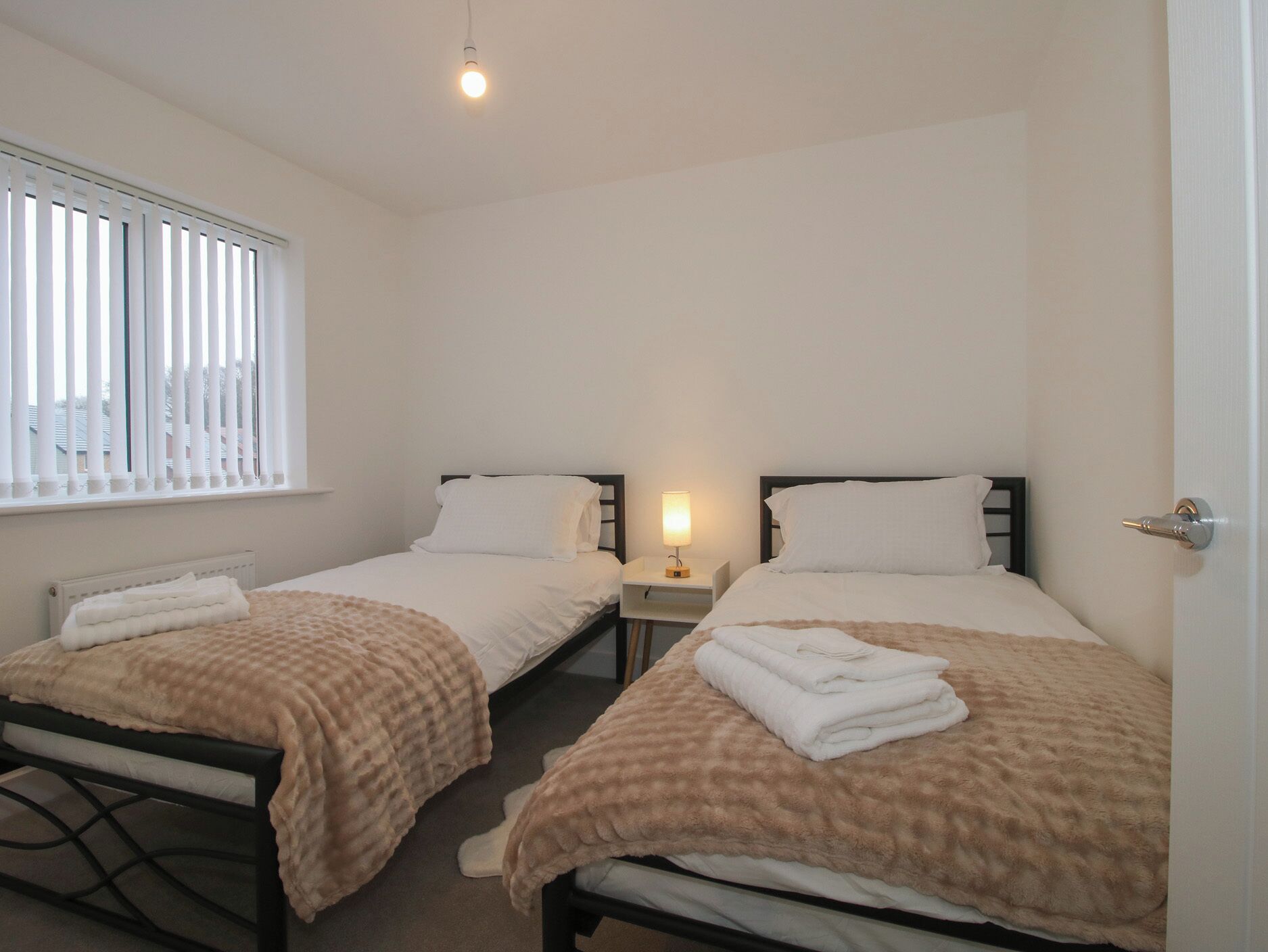 Une chambre double avec deux lits, chacun doté d’une table de chevet et d’une lampe, une fenêtre avec des stores et un radiateur placé sous la fenêtre.