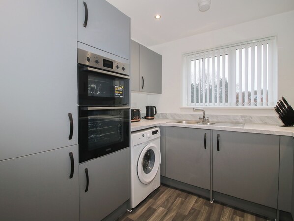 A modern kitchen with a built-in oven, a washing machine, a sink, and a window with blinds.