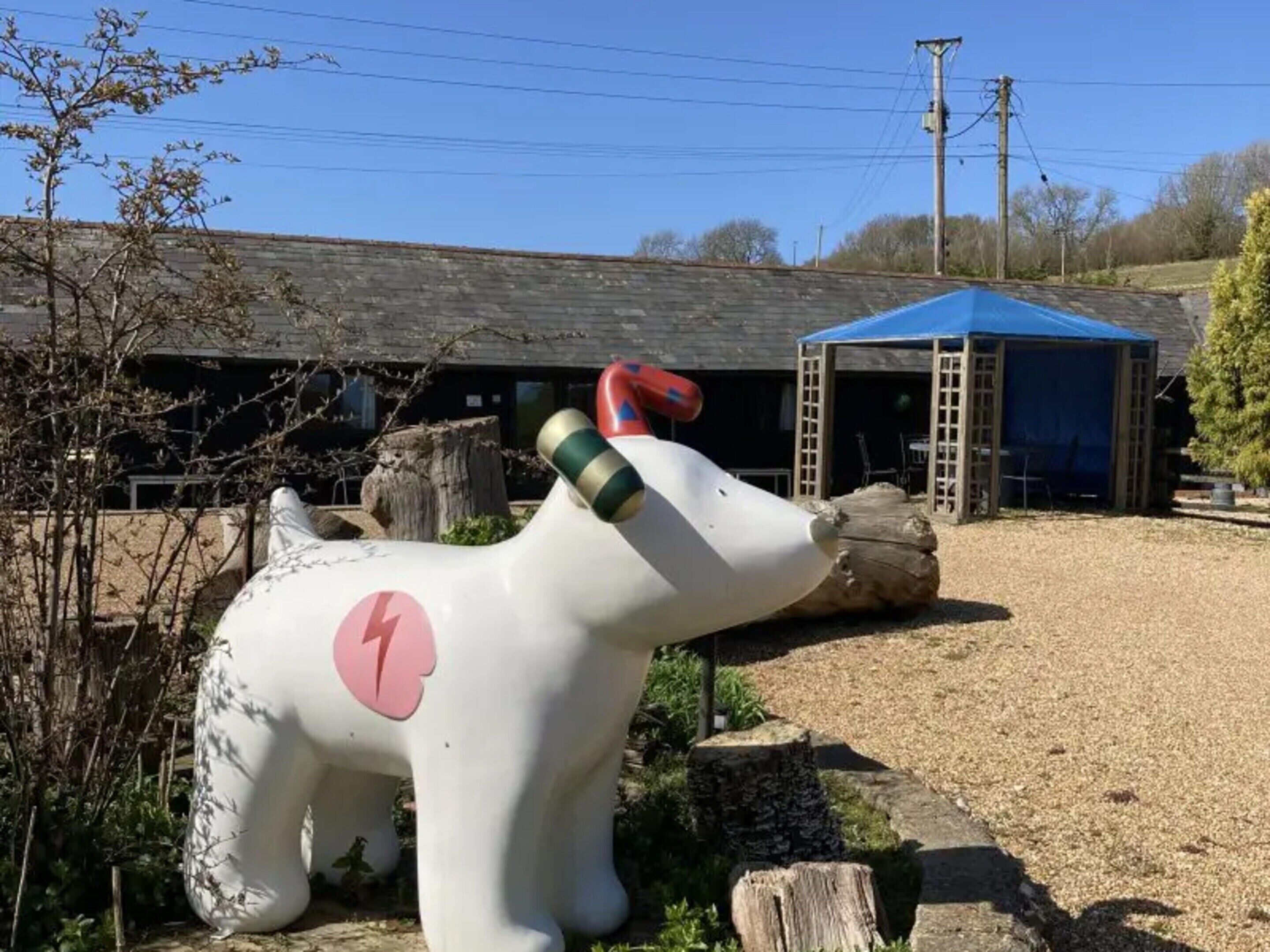 A large white sculpture of a dog with a pink heart on its side, positioned in front of a building with a blue roof and a wooden structure nearby.