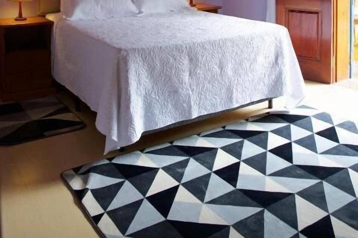 Ein Schlafzimmer mit einem Bett, zwei Nachttischlampen, einer Holzbalkendecke und einem Teppich mit geometrischem Muster.