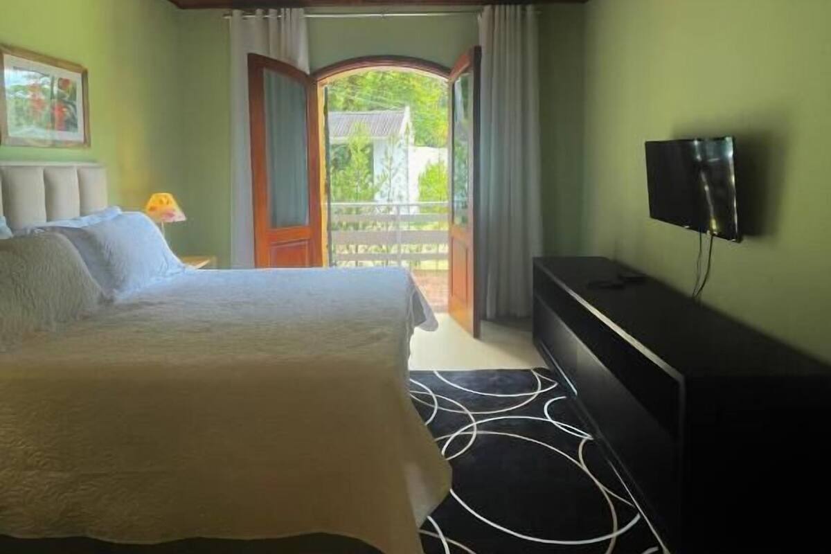 Ein Schlafzimmer mit Holzboden, einem Bett, einem Fernseher und Blick auf einen Garten durch eine offene Tür.