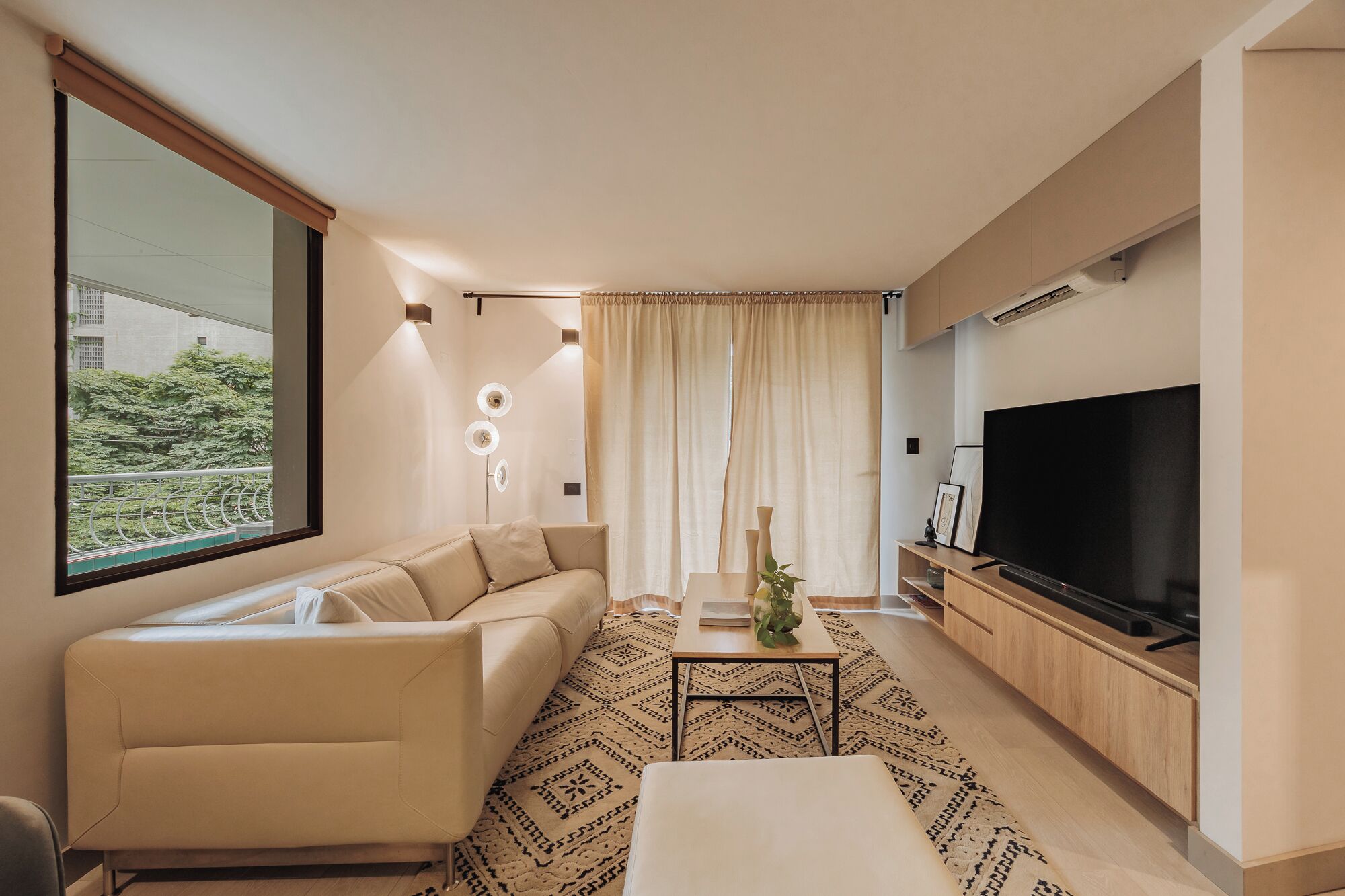 En moderne stue med sofa, sofabord, fjernsyn og et stort vindue med udsigt over grønne områder.
