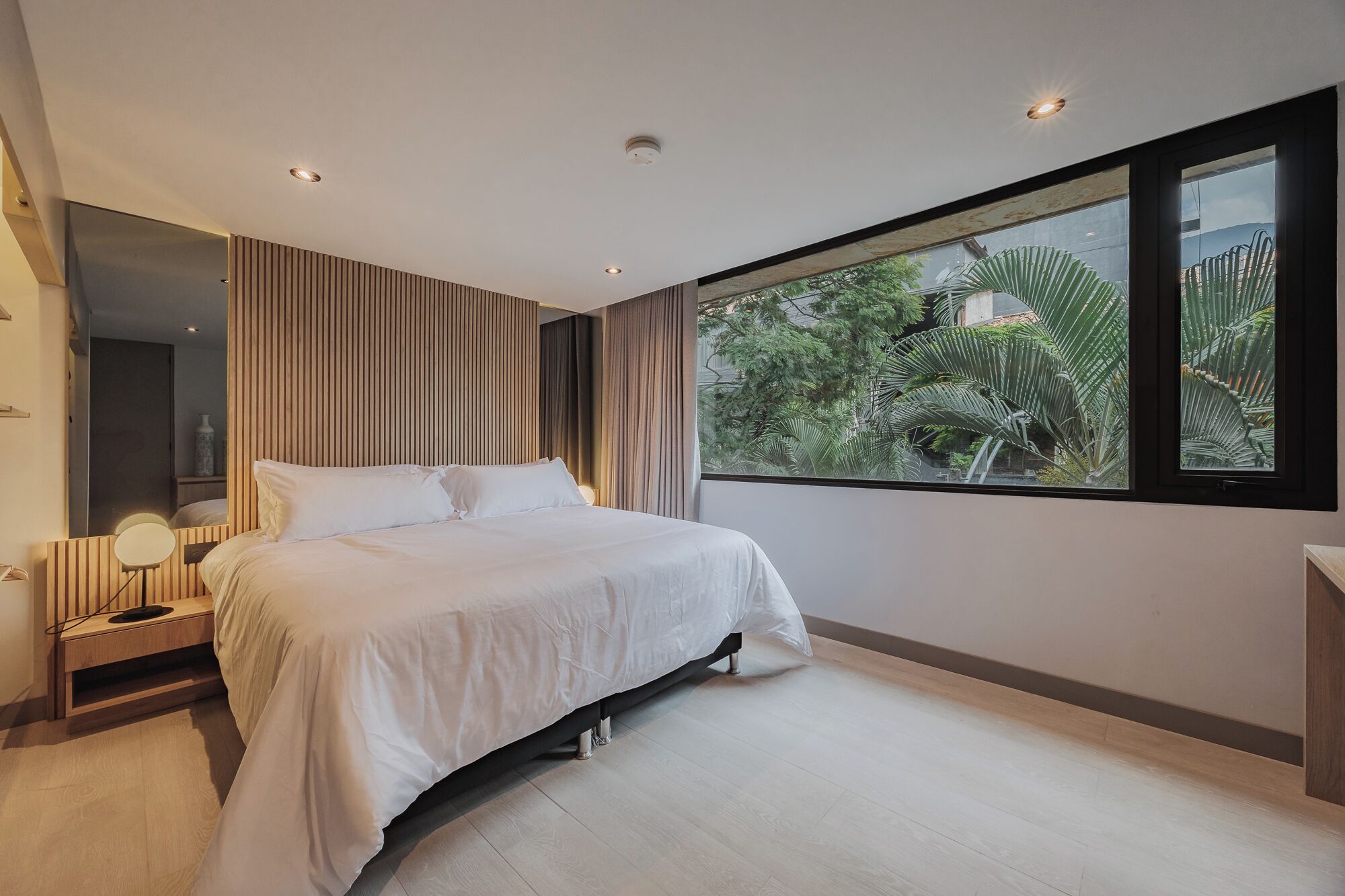 A modern bedroom with a large bed, a bedside table with a lamp, a mirror, and a window with a view of greenery.