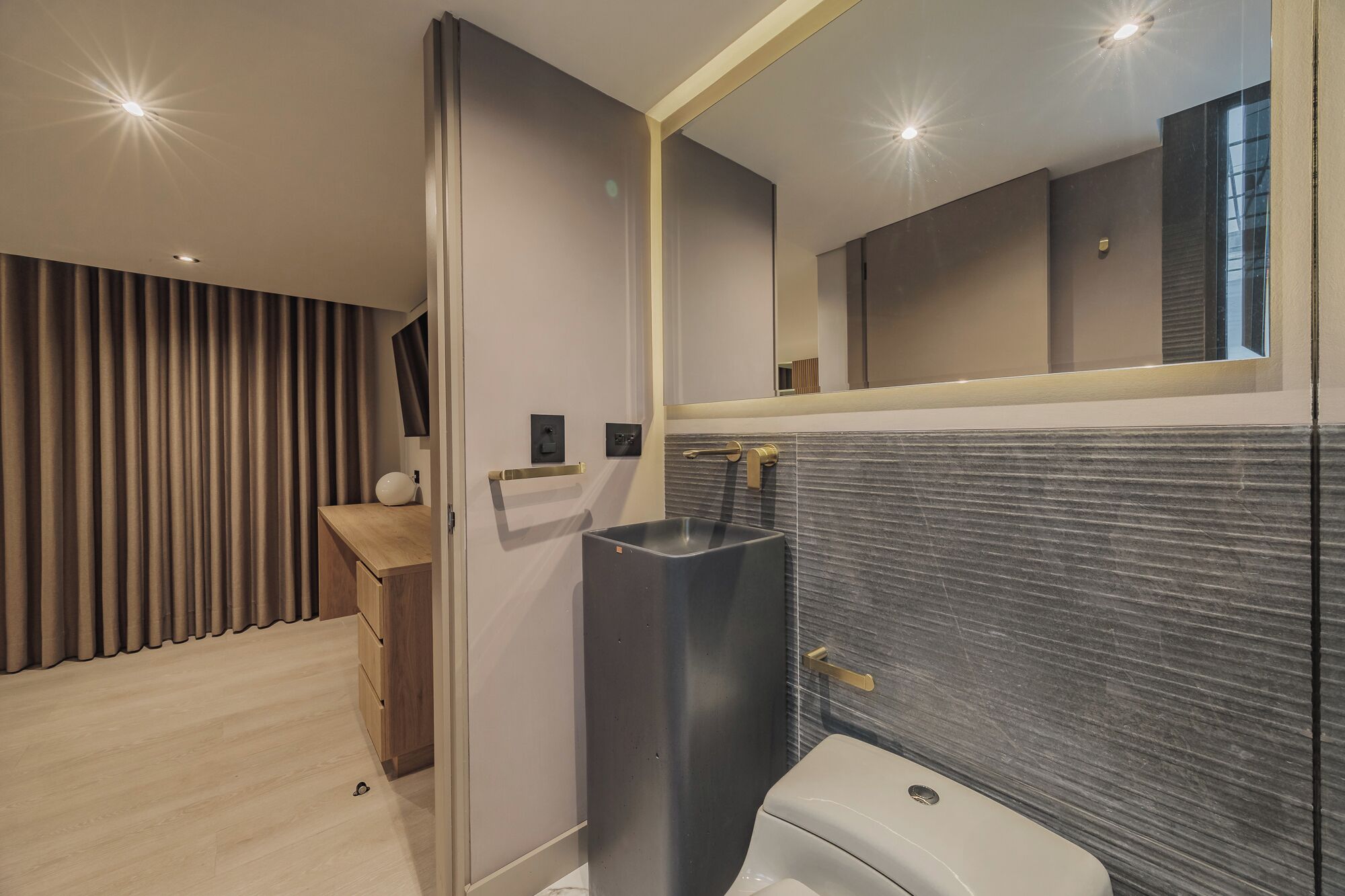 A modern bathroom with a dark tiled wall, a large mirror, and a wooden vanity.