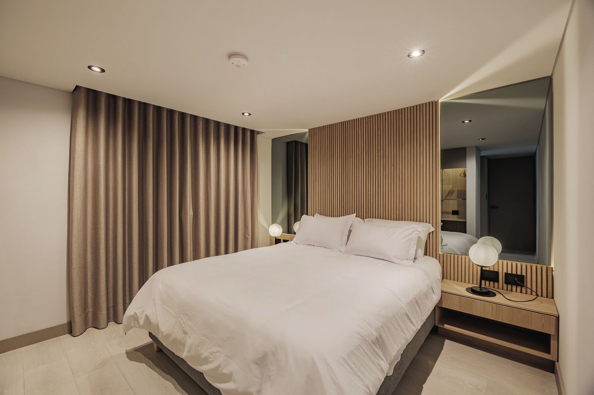 Ein modernes Hotelzimmer mit einem großen Bett, einem Nachttisch mit Lampe, einem Spiegel und Blick auf die Stadt durch das Fenster.