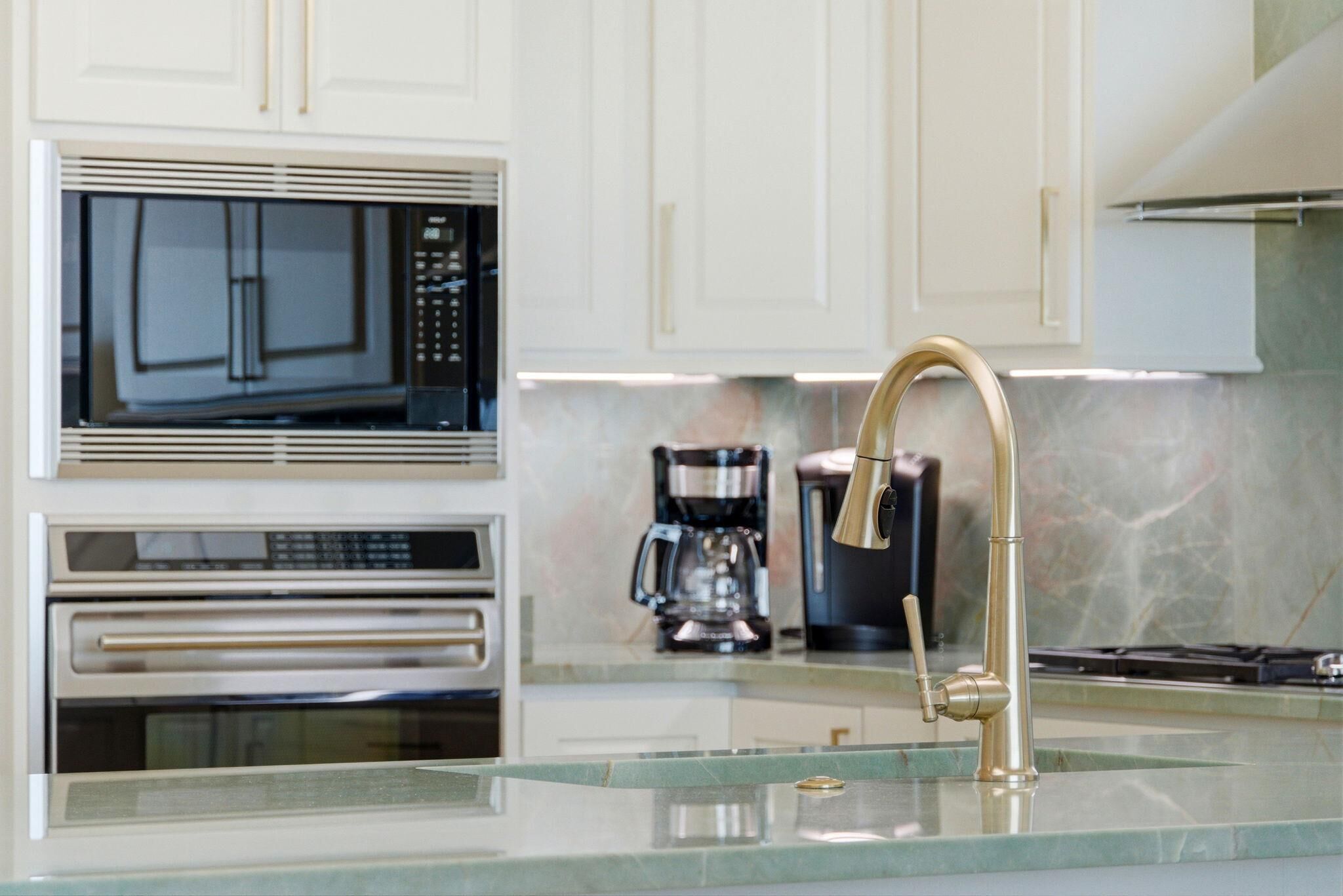 A modern kitchen with white cabinetry, a stainless steel oven, a microwave, a coffee maker, and a gold faucet.