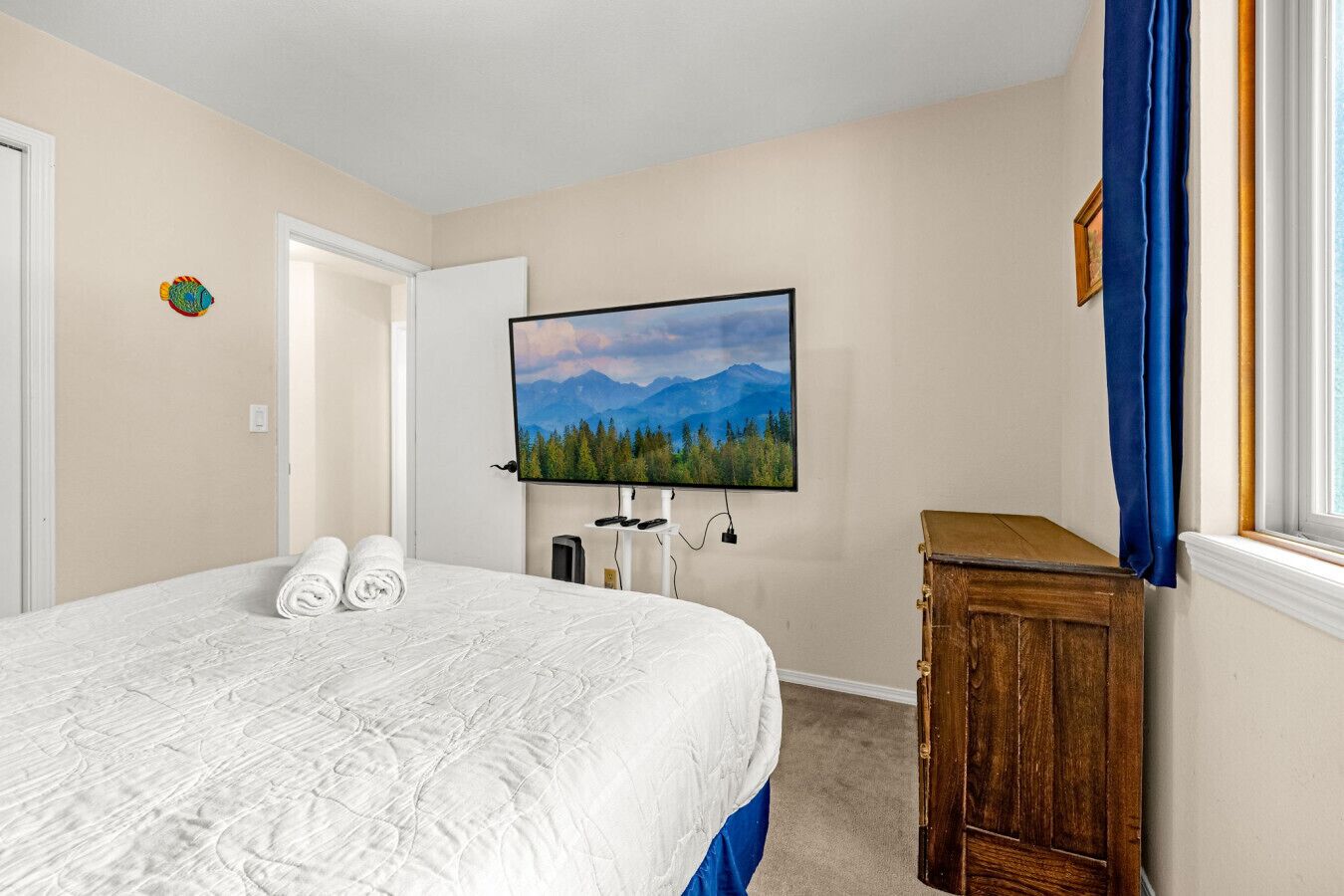 A hotel room with a bed, a television mounted on the wall, a wooden dresser, and a window with blue curtains.
