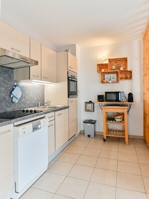 A modern kitchen with light wood cabinets, a built-in oven, a microwave, and a wooden shelf with decorative items. - Côte Fleurie · Cosy Norman apt, balcony & parking (Saint-Arnoult)
