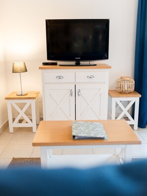 A modern room with a flat-screen TV on a wooden stand, a white cabinet with two drawers, a small table with a cushion, a lamp, and a decorative wooden bowl. - Côte Fleurie · Cosy Norman apt, balcony & parking (Saint-Arnoult)