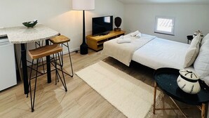 Een kleine slaapkamer met een bed, nachtkastje, lamp en een klein kitchenettegedeelte met een tafel en krukjes.