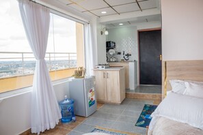 Une chambre d’hôtel avec un lit, une petite kitchenette, un distributeur d’eau et une vue sur la ville.