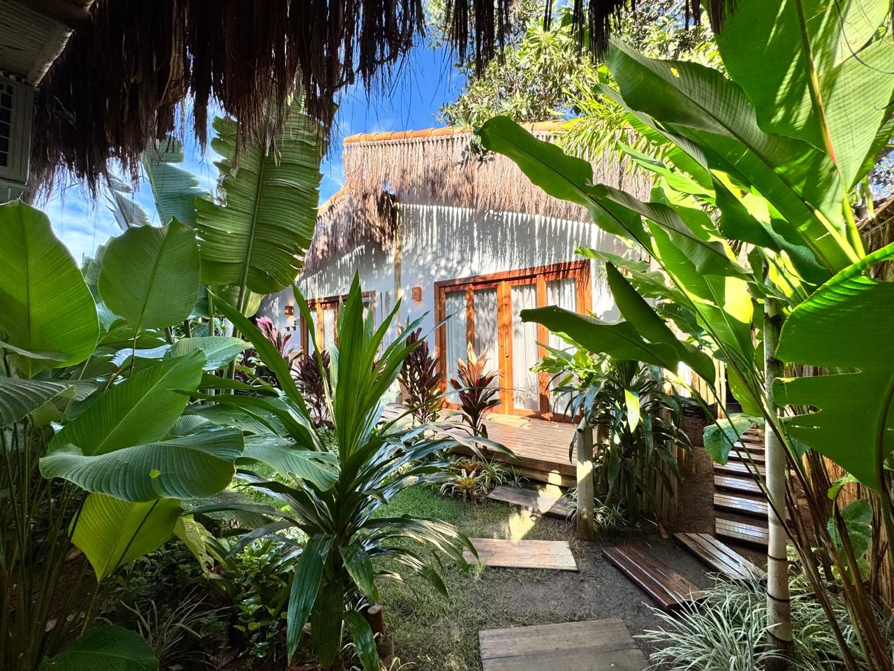 Un jardin tropical doté d’une terrasse en bois, d’une structure à toit de chaume et d’une végétation luxuriante.