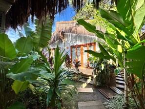 A tropical garden with a wooden deck, a thatched-roof structure, and lush greenery. - Casa Preguiça (Corumbau)