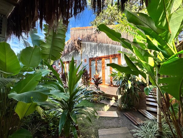 A tropical garden with a wooden deck, a thatched-roof structure, and lush greenery. - Casa PreguiƧa (Corumbau)
